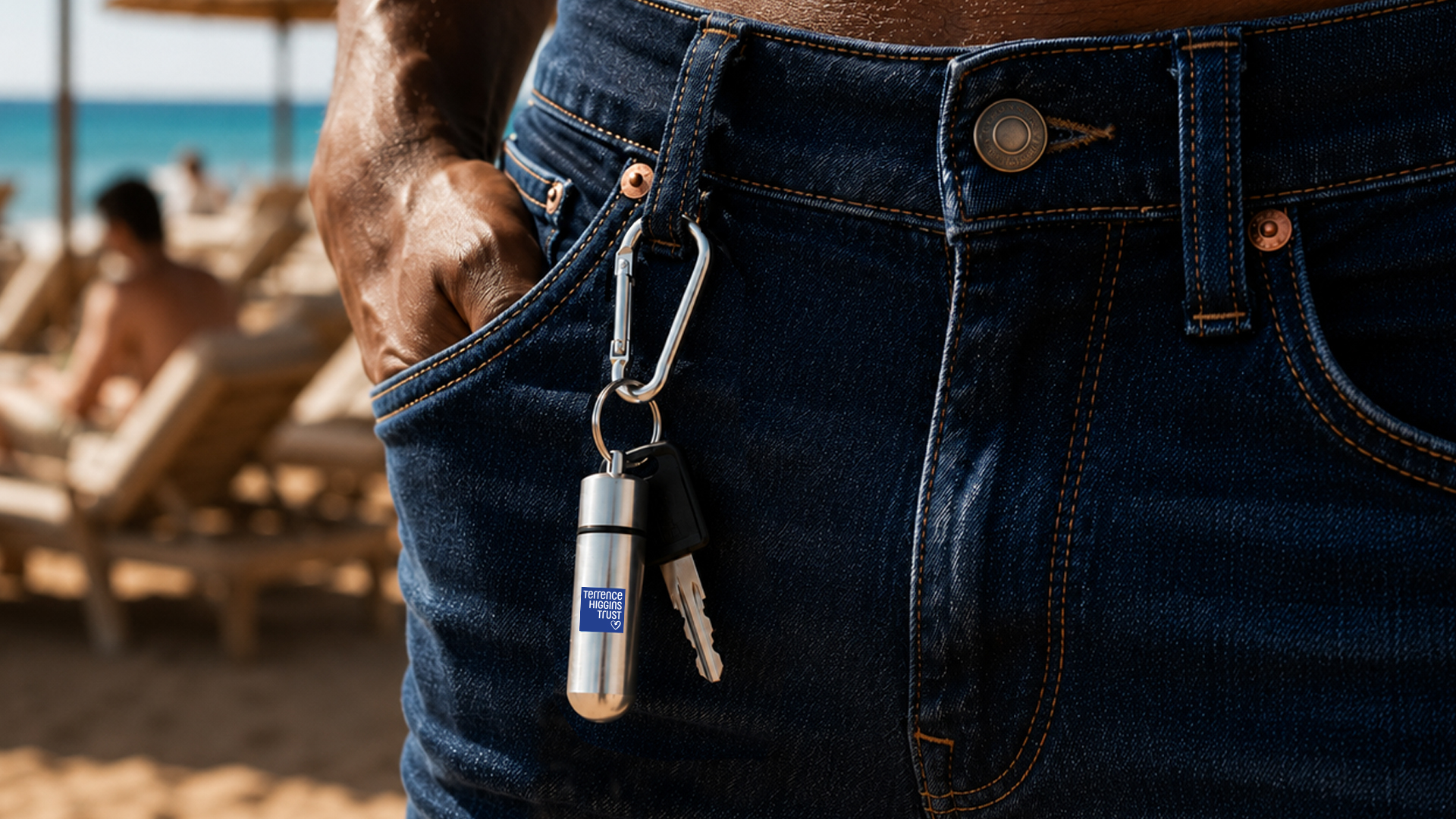 A person is standing on a beach with their hand in their jeans pocket. A silver pill holder keyring with the Terrence Higgins Trust logo hangs off a belt loop.
