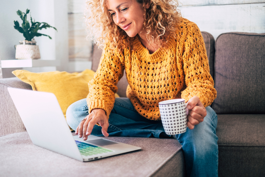 A woman in a yellow jumper looks at her laptop, holding a mug.