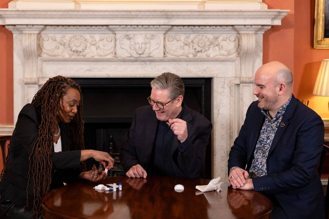 Beverley Knight, Keir Starmer and Richard Angell sit at a table in front of fireplace, performing an HIV test.