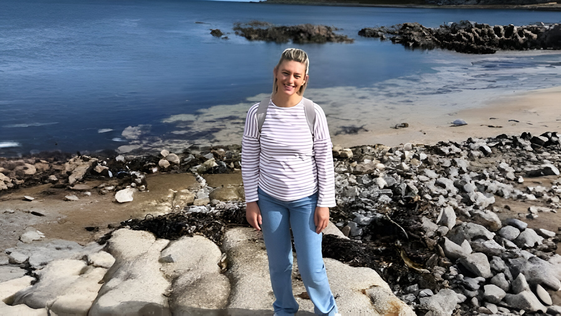 A white woman in a white striped top standing on a rocky beach.