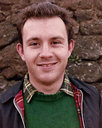 Image from the 1980s showing Mark Asthon. He is has brown hair and a green jumper and he is smiling at the camera.