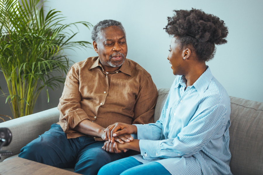 An older man sits on a sofa holding hands and talking to a woman.