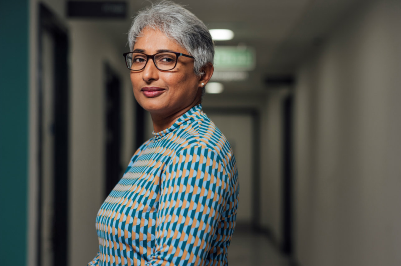 A portrait of a mid adult, South Asian female, standing in a low lit corridor, wearing glasses. This is a model used to represent Priya.