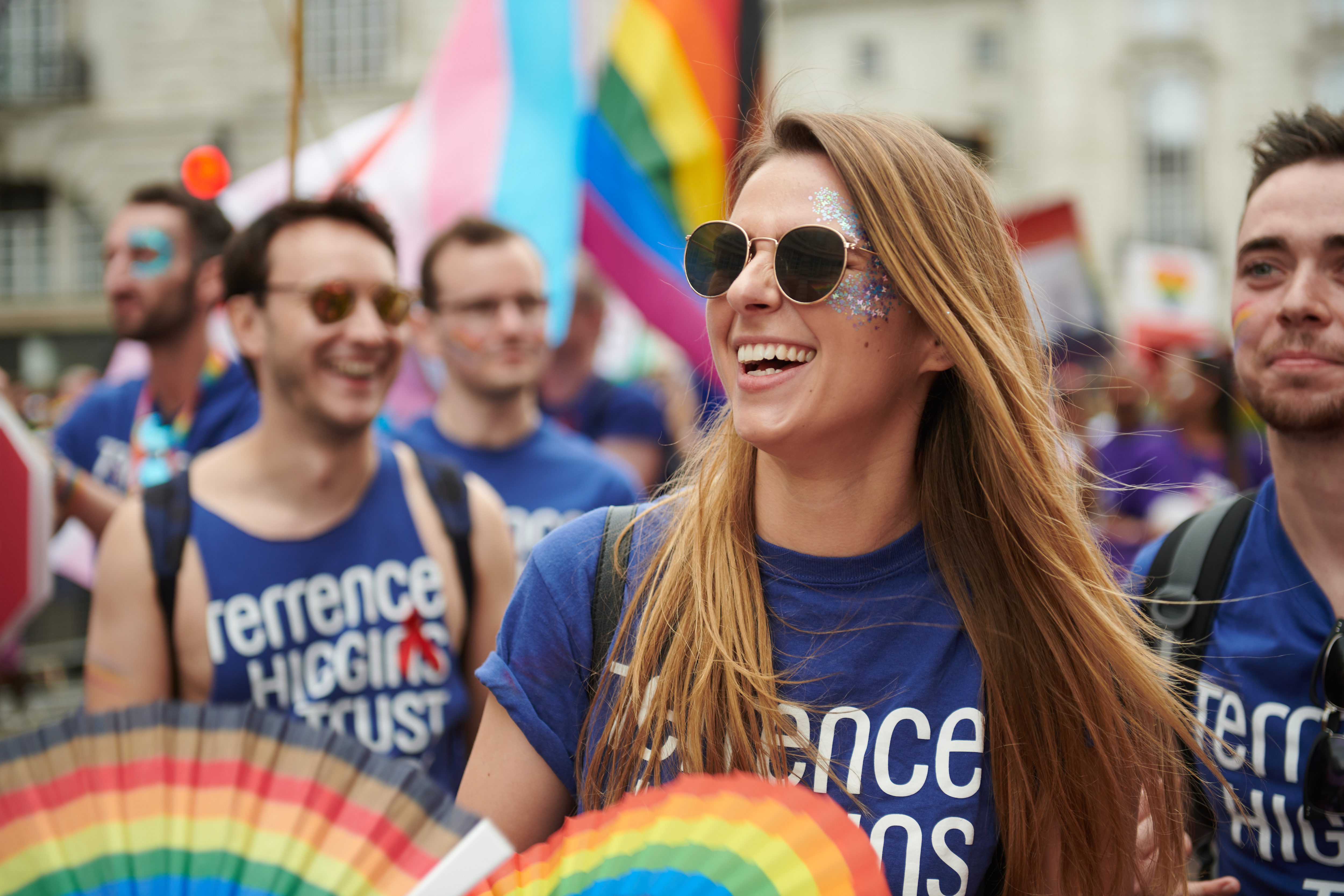 A group of people at a Pride march. They're wearing Terrence Higgins Trust blue t-shirts and carrying rainbow fans.