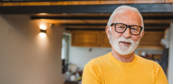 An older man wearing a yellow jumper looks at the camera.