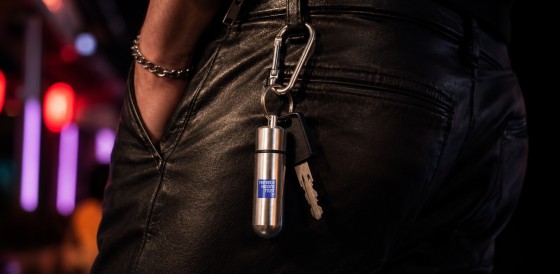 Close-up of a silver capsule keyring with the Terrence Higgins Trust logo attached to a carabiner and key, clipped to the waistband of black leather trousers. The background features colourful blurred lights.