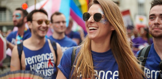 A group of people at a Pride march. They're wearing Terrence Higgins Trust blue t-shirts and carrying rainbow fans.
