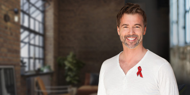 A smiling man in a white shirt with a red ribbon pinned to it. The background shows a loft apartment.