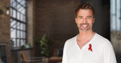 A smiling man in a white shirt with a red ribbon pinned to it. The background shows a loft apartment.