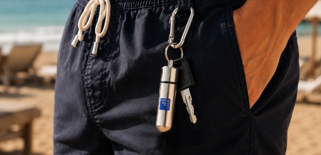 Close-up of a person wearing navy shorts, standing on a beach. A silver keyring with the THT logo on it, hangs off the shorts.