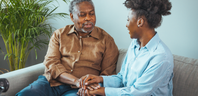 An older man sits on a sofa holding hands and talking to a woman.
