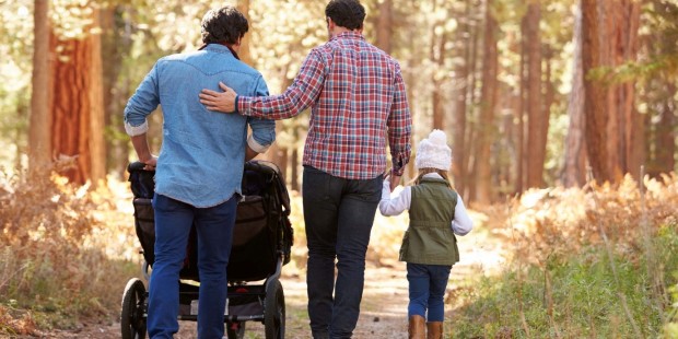 A same sex parent family walks their daughter and infant in a pushchair for walk in the woods.