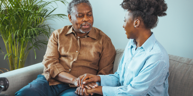 An older man sits on a sofa holding hands and talking to a woman.