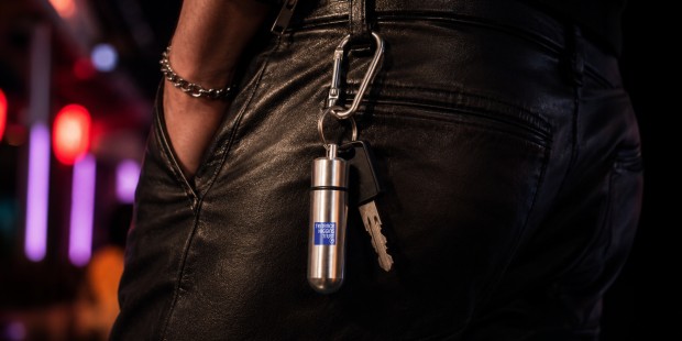 Close-up of a silver capsule keyring with the Terrence Higgins Trust logo attached to a carabiner and key, clipped to the waistband of black leather trousers. The background features colourful blurred lights.