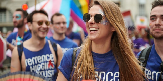 A group of people at a Pride march. They're wearing Terrence Higgins Trust blue t-shirts and carrying rainbow fans.