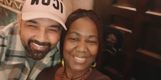 Two friends embracing. The man has his arm around the woman and they are smiling at the camera.